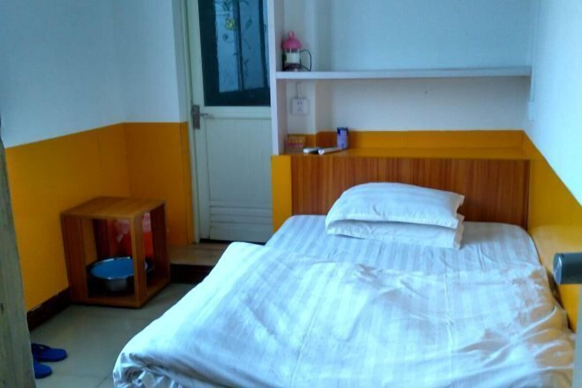 A small, single-bed room with a wooden headboard, a bedside table, and a shelf above the bed.