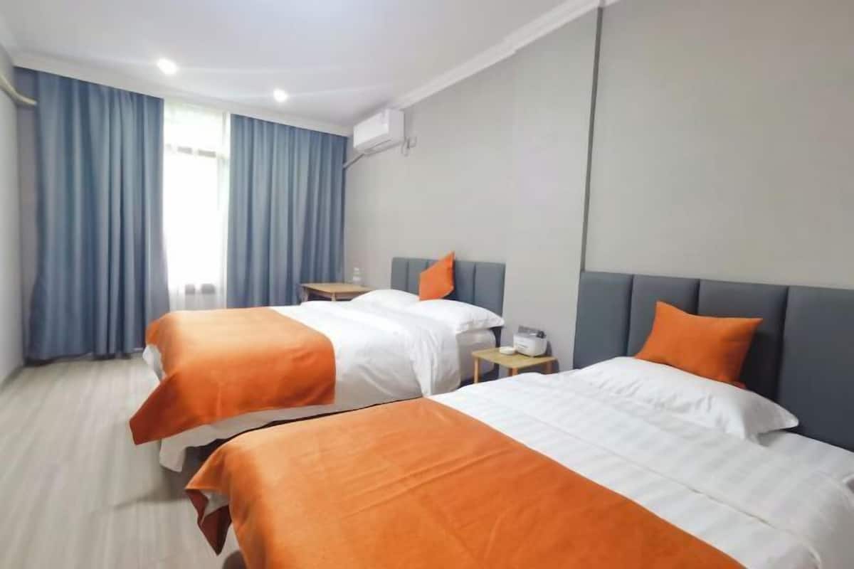 Une chambre d’hôtel avec deux lits, chacun recouvert d’une couverture orange, une table de chevet avec une lampe et une fenêtre avec des rideaux bleus.