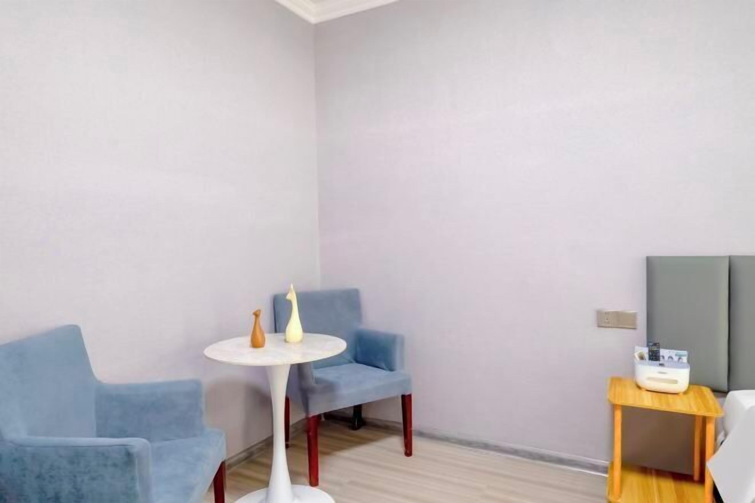 A room with a blue chair, a small round table, a bedside table with a lamp, and a bed.