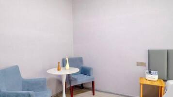 A room with a blue chair, a small round table, a bedside table with a lamp, and a bed.
