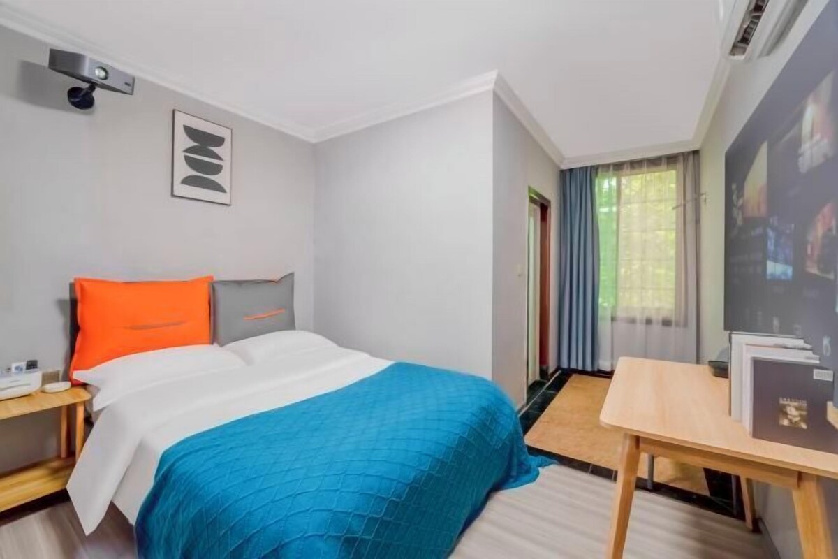 A hotel room with a bed, a desk, a chair, a window with curtains, and a wall-mounted projector.
