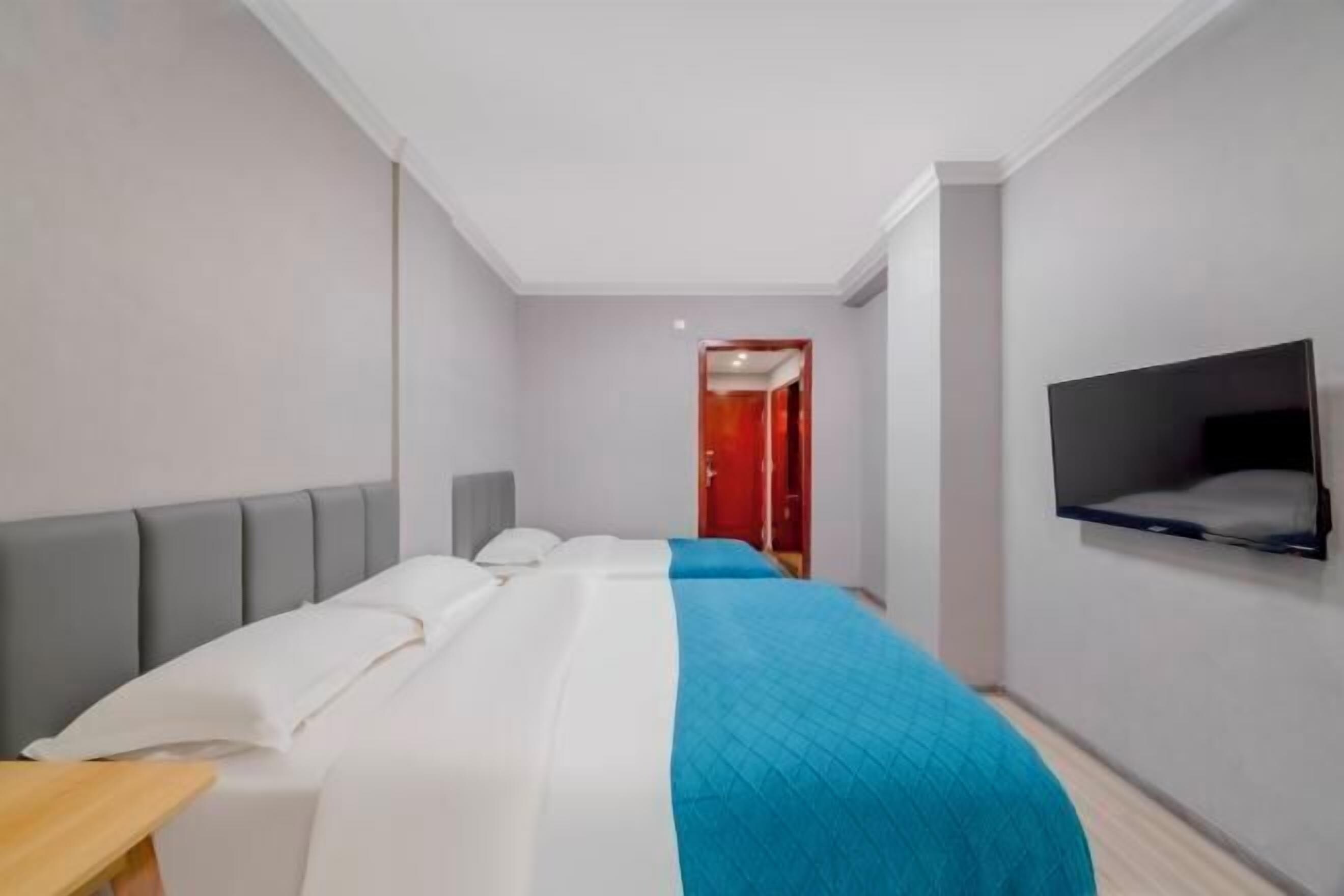 A modern hotel room with a bed, a TV mounted on the wall, and a red door in the background.
