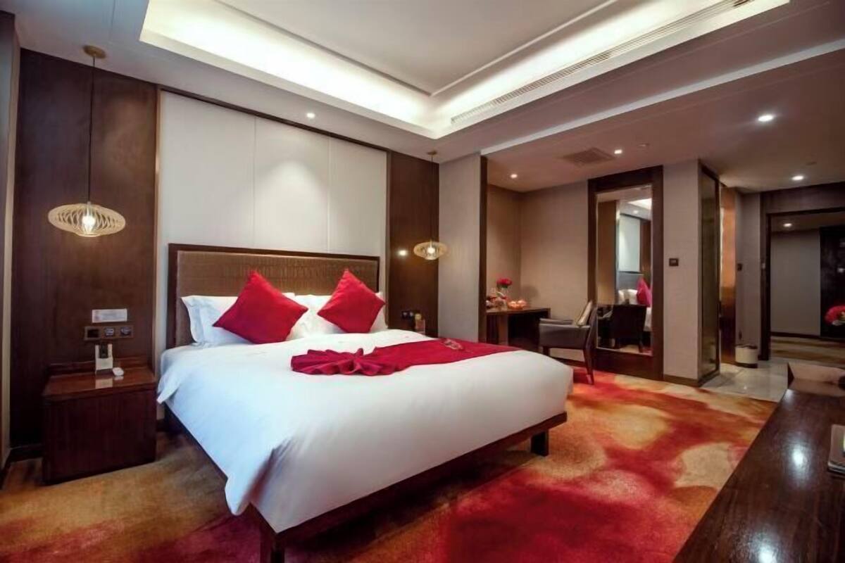 A hotel room with a large bed, red pillows, a wooden headboard, a bedside table with a lamp, and a carpeted floor.