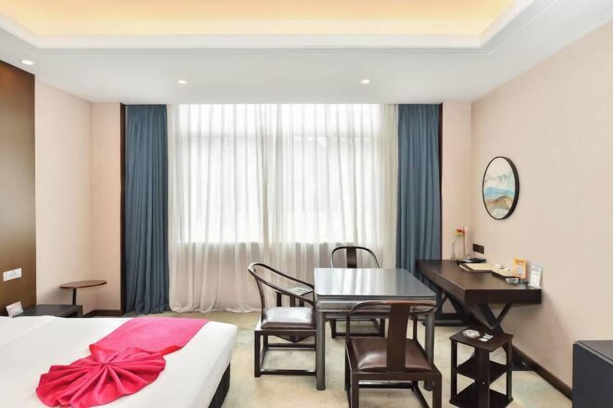 A hotel room with a bed, a desk, two chairs, a small table, and a large window with curtains.