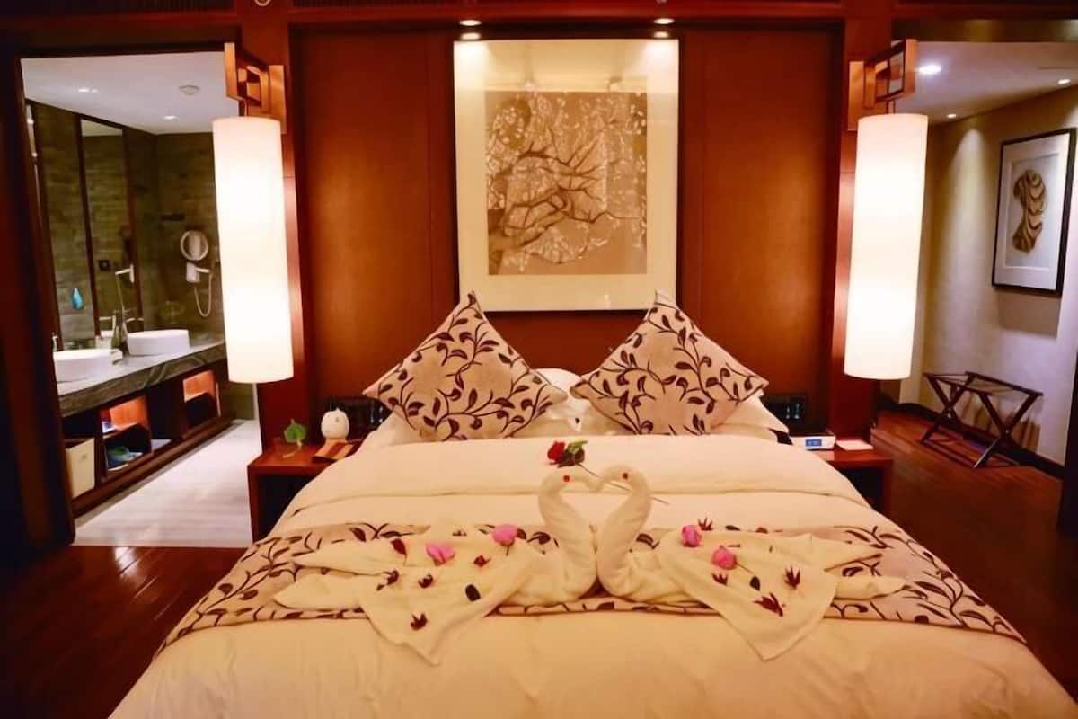 Quarto de hotel com uma cama grande, duas mesas de cabeceira, um espelho, banheiro com pia e chuveiro, e um quadro na parede.