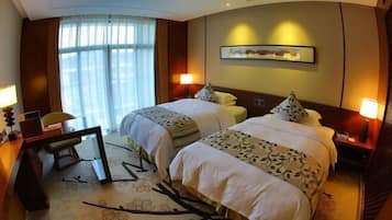 A hotel room with two beds, a desk, a chair, a large window with curtains, and a framed picture on the wall.