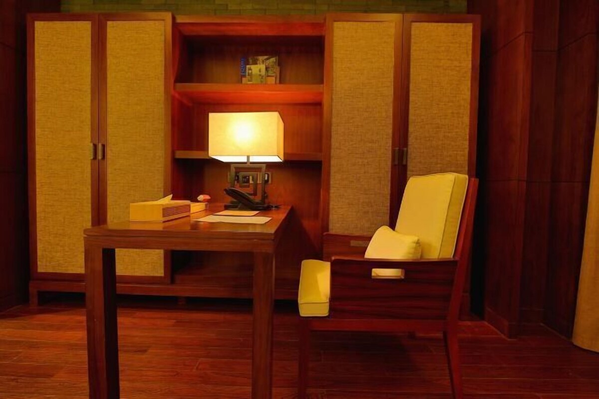 A room with a wooden desk, a chair, a lamp, and a shelf with books and a box.