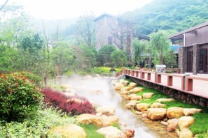 A hot spring area with a stone pathway, lush greenery, and a building in the background. - Chuishan Hot Spring Resort (Zhenjiang)