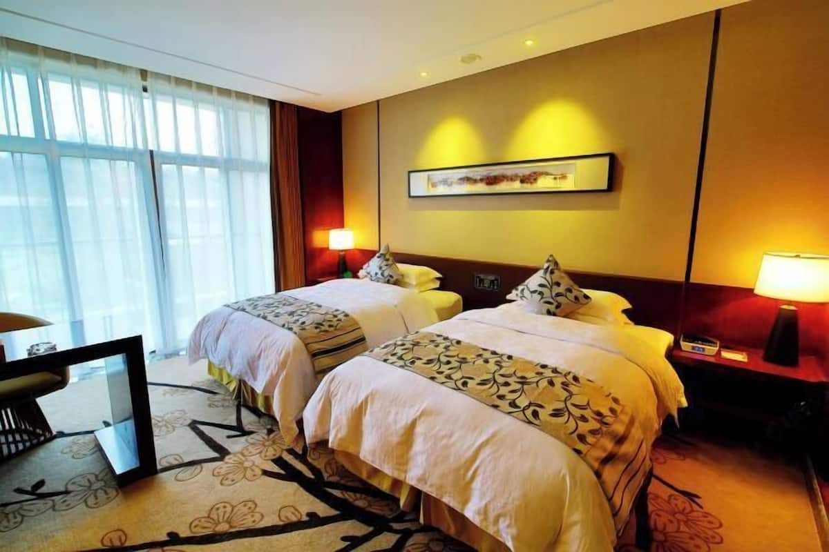 Quarto de hotel com duas camas, uma escrivaninha e uma cadeira. Possui uma janela grande com cortinas, um quadro na parede e duas luminárias de cabeceira.