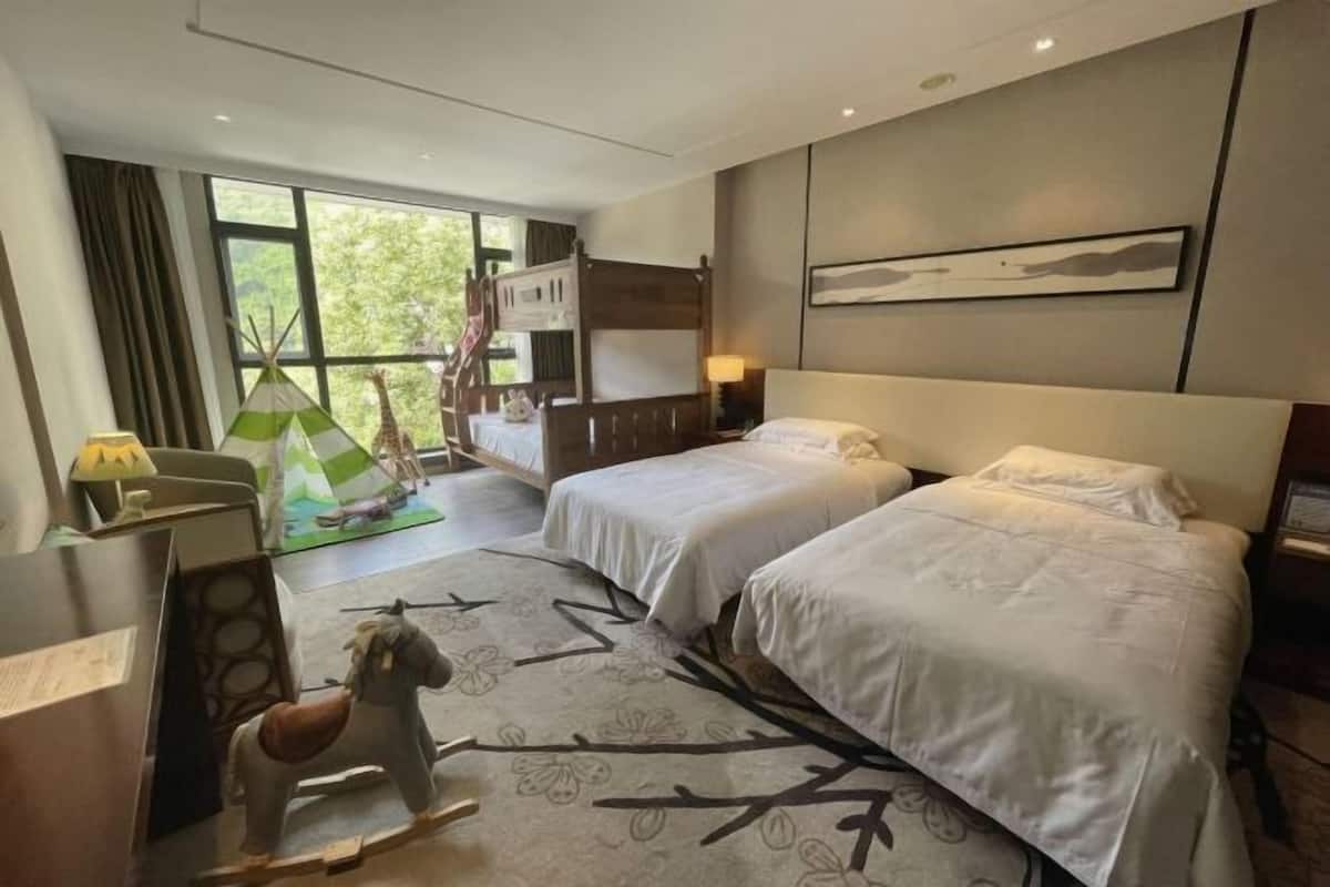 Quarto de hotel com duas camas, uma janela grande, vista para o verde lá fora, uma pequena área de recreação com uma girafa e um cavalo, e um quadro na parede.
