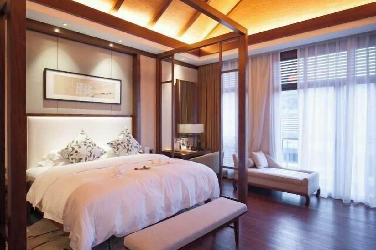 Quarto com cama grande, mesa de cabeceira, banqueta e vista para o exterior através de cortinas.