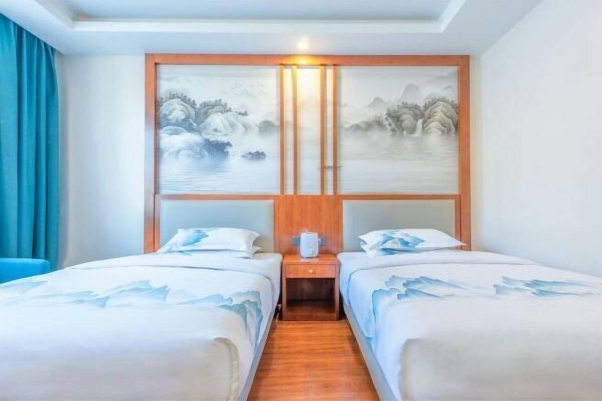 A hotel room with two beds, a wooden headboard, a painting of a mountainous landscape, and a nightstand with a lamp.