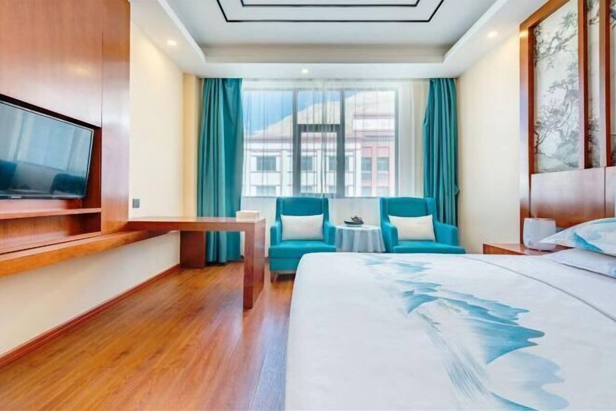 A hotel room with a large bed, a TV mounted on the wall, a small table, two chairs, and a window with curtains.