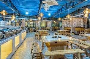 A restaurant with a high ceiling, exposed beams, and a blue tarp covering the top. There are wooden tables and benches, and a buffet area with stainless steel containers.