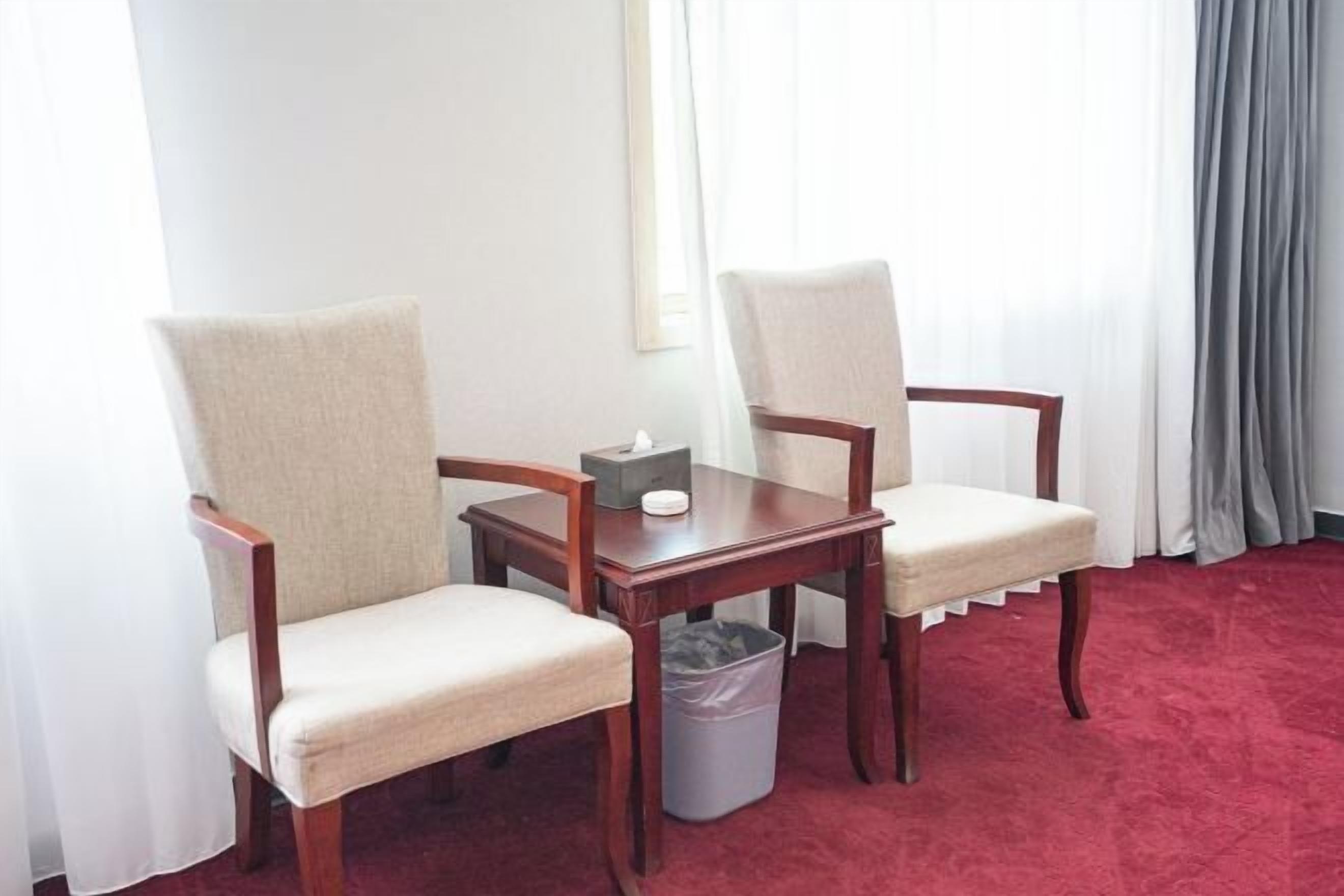 A hotel room with two upholstered chairs, a wooden table, a trash bin, and a red carpet.