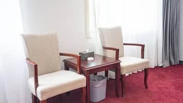 A hotel room with two upholstered chairs, a wooden table, a trash bin, and a red carpet.