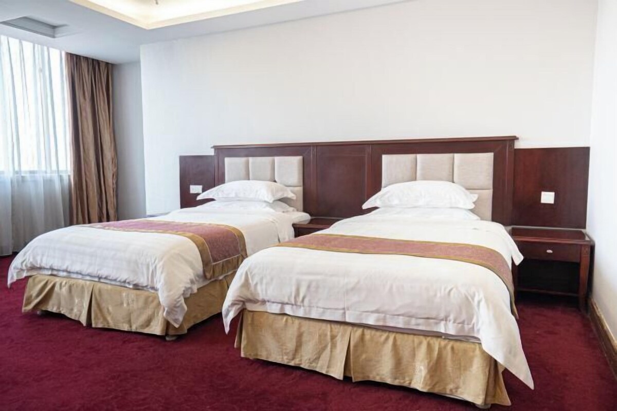 A hotel room with two beds, a wooden headboard, a red carpet, and a window with curtains.