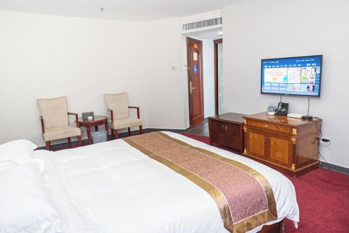 A hotel room with a bed, two chairs, a wooden cabinet, and a television displaying a menu.