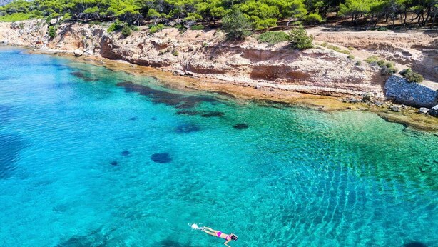 Une mer d’un bleu profond où une personne nage, entourée d’un paysage rocheux et d’arbres verdoyants.