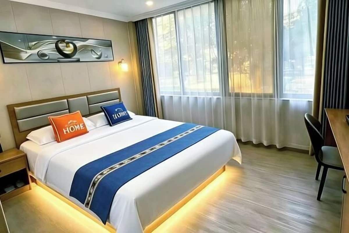 A hotel room with a bed, a desk, a chair, a large window with curtains, and a wall-mounted artwork.