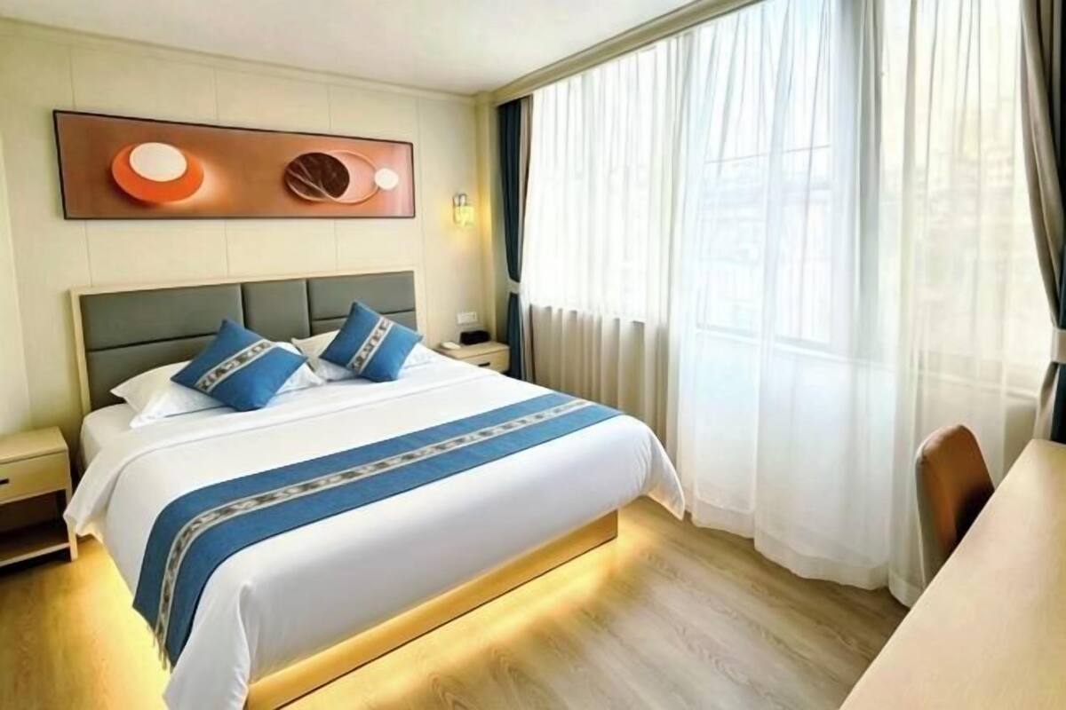 A hotel room with a large bed, a bedside table, a nightstand, a wall-mounted light, and a window with curtains.