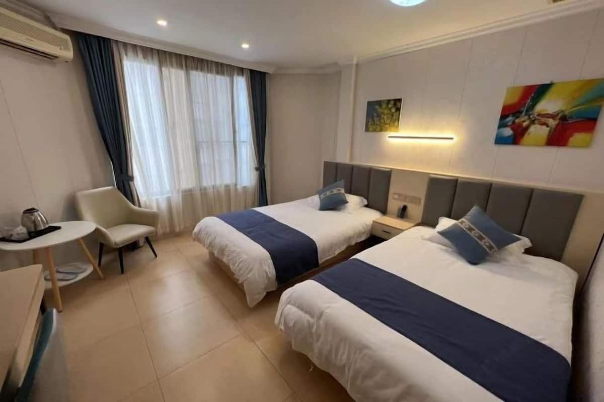 A hotel room with two beds, a small table, a chair, a desk, an air conditioning unit, and a window with curtains.