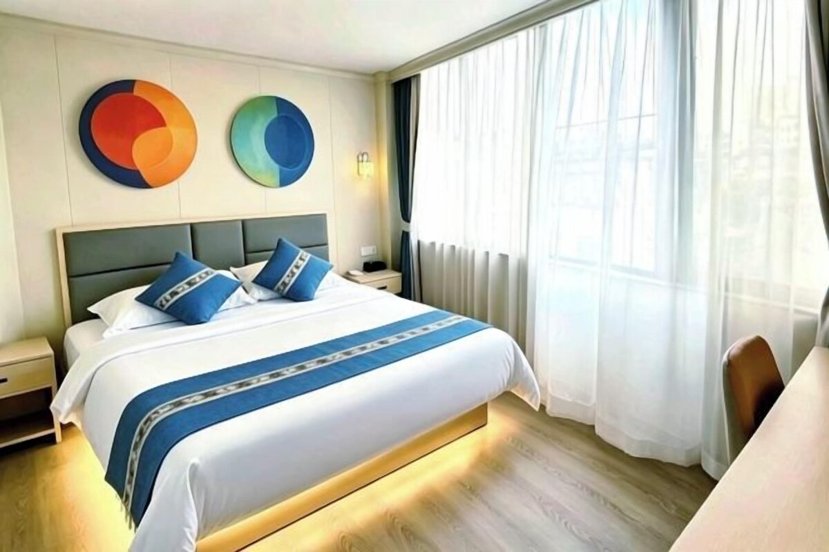 Une chambre d’hôtel avec un lit, une table de chevet, une lampe, une chaise et une fenêtre avec des rideaux.