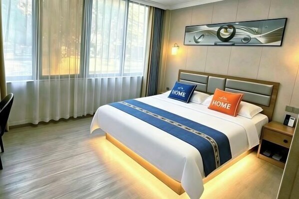 A hotel room with a bed, a nightstand, a chair, a window with curtains, and a wall-mounted artwork.