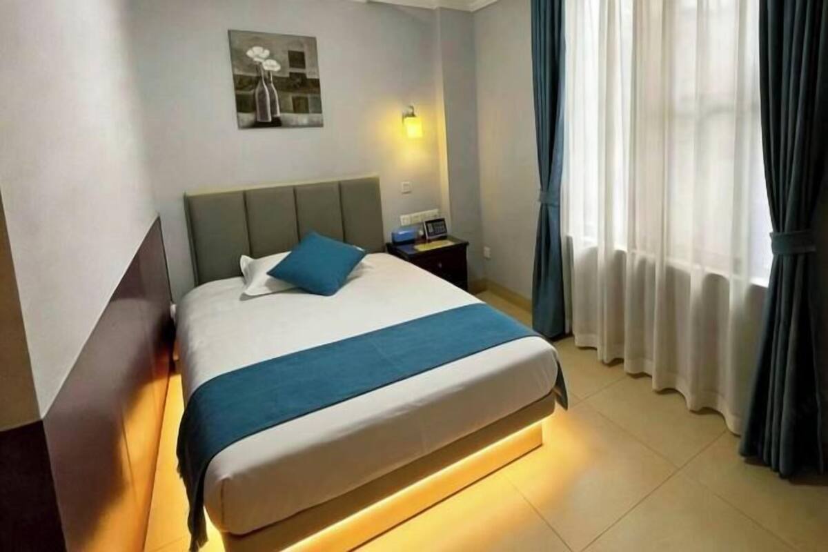 A hotel room with a bed, a nightstand, a wall-mounted light, and a window with curtains.