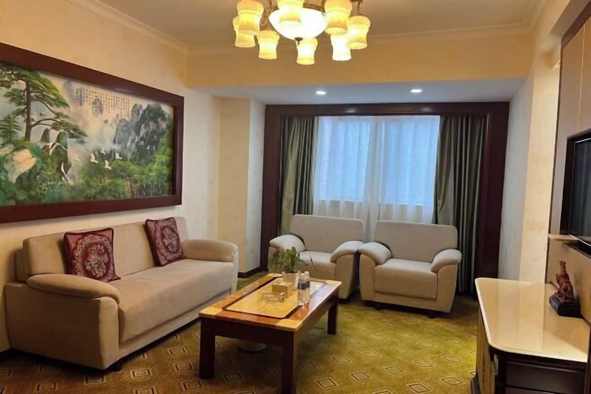A hotel room with a beige sofa, two armchairs, a wooden coffee table, and a painting of a forest scene.