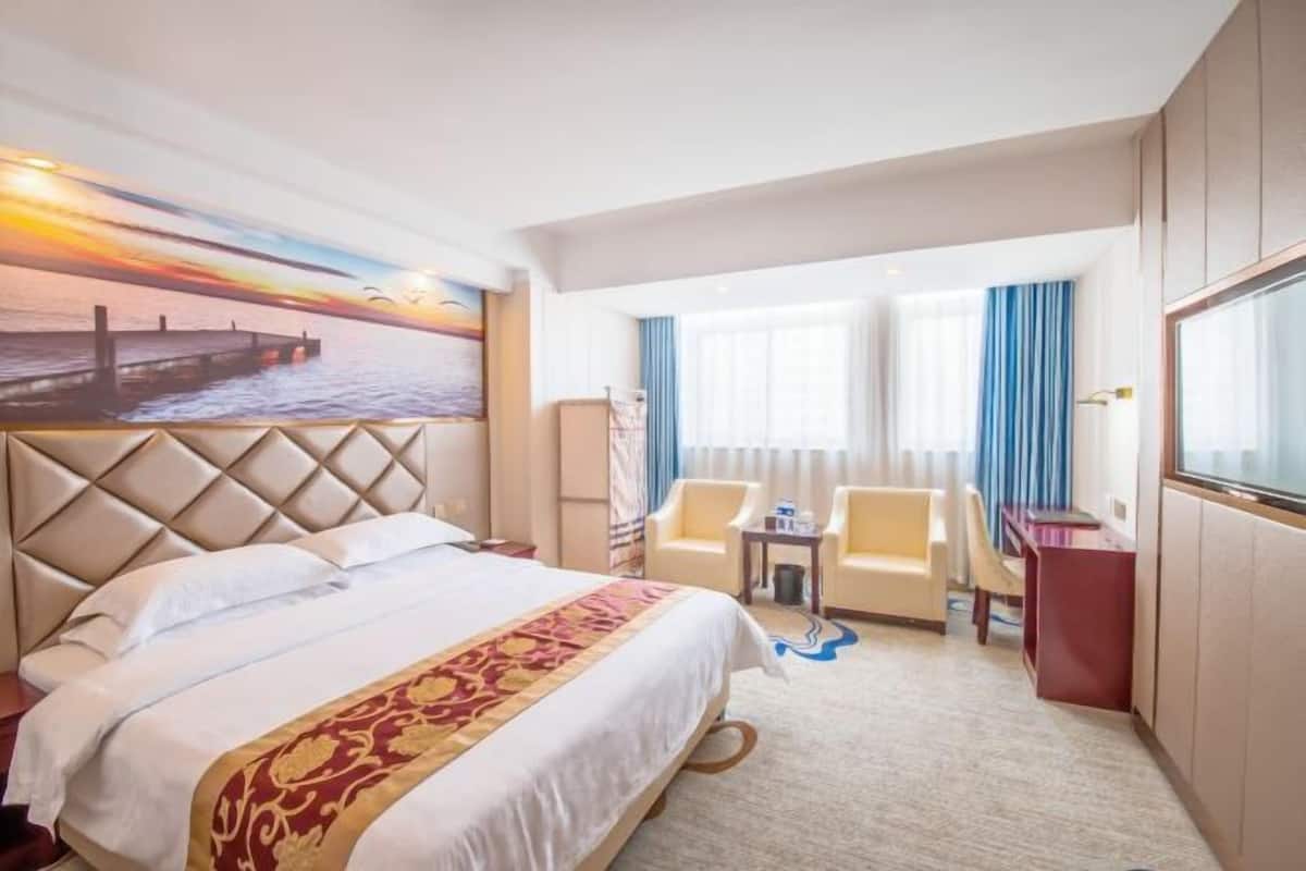A hotel room with a large bed, a desk, two chairs, a small table, and a painting of a sunset over a pier.