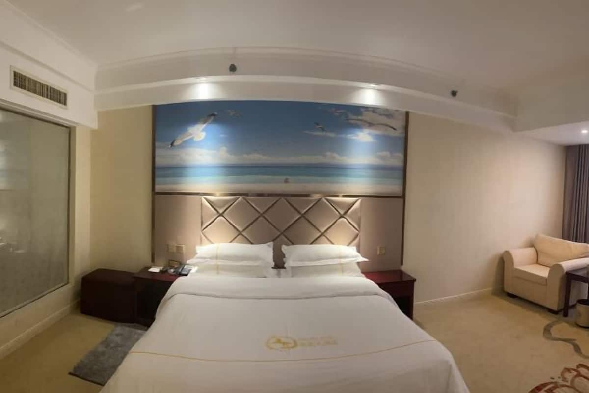A hotel room with a large bed, a headboard with a sky and clouds mural, a bedside table, a chair, and a view of the sea through a glass door.