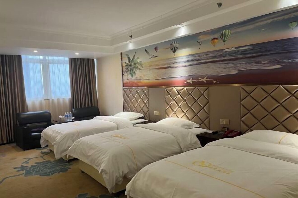 A hotel room with three beds, a large painting of a beach scene, and a view of the ocean through the window.