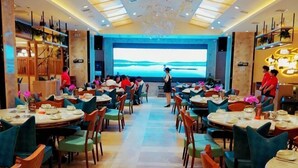Un restaurante con varias mesas y sillas, una pantalla grande que muestra un paisaje pintoresco y una decoración interior moderna.