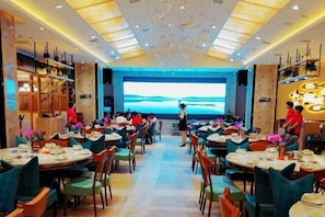 Un restaurante con varias mesas y sillas, una pantalla grande que muestra un paisaje pintoresco y una decoración interior moderna.