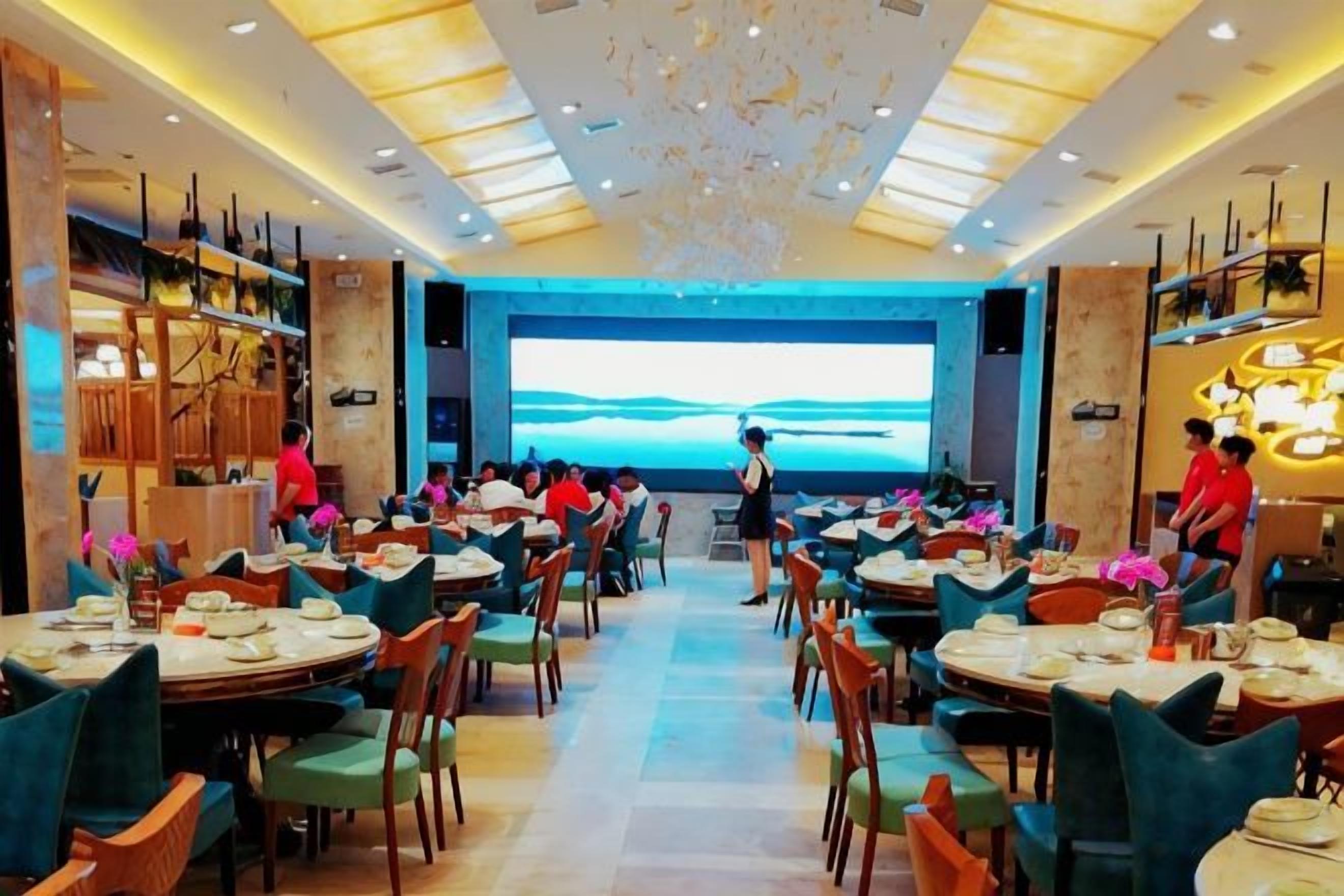 A restaurant with multiple dining tables and chairs, a large screen displaying a scenic view, and a modern ceiling design.