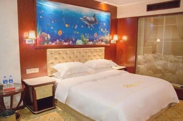 A hotel room with a large bed, a nightstand, a small table, and a wall painting of an underwater scene. - Xing mei da Hotel (Changsha County)