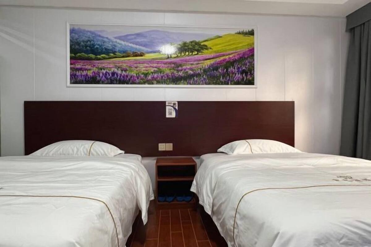 A hotel room with two beds, a wooden headboard, a painting of a landscape, and a small shelf with shoes.
