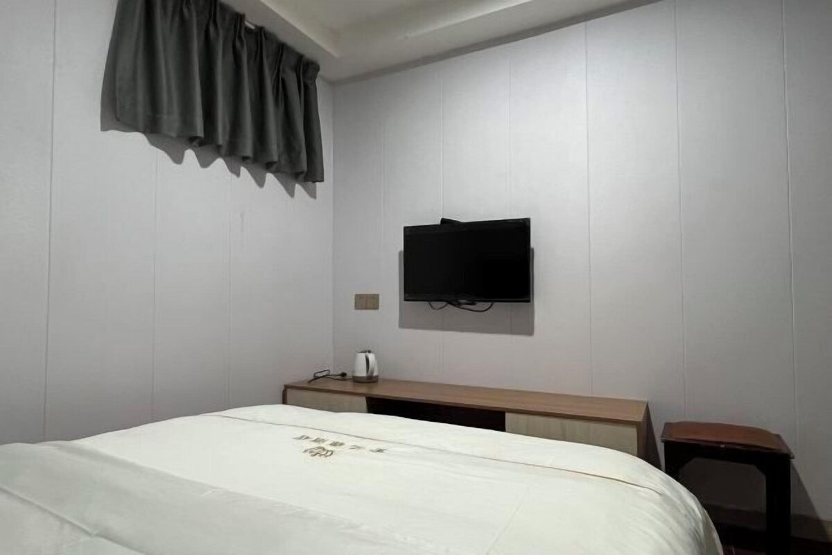 A hotel room with a bed, a flat-screen TV mounted on the wall, a bedside table with a kettle, and a small wooden stool.