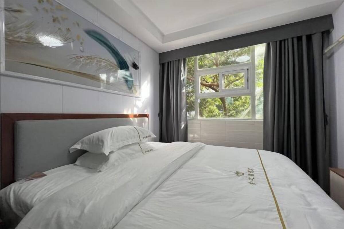 A neatly made bed with white linens and pillows, a wooden headboard, and a window with curtains.