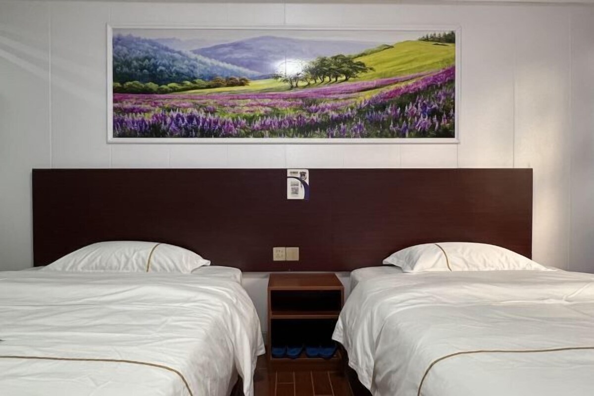 A hotel room with two beds, a headboard with a painting of a landscape, and a small wooden nightstand with a blue towel.