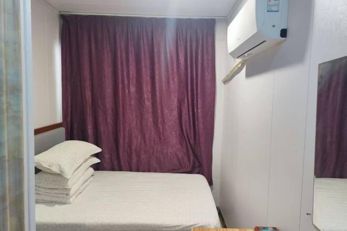 A small, compact room with a bed, a stack of white pillows, a window with purple curtains, and a wall-mounted air conditioning unit.