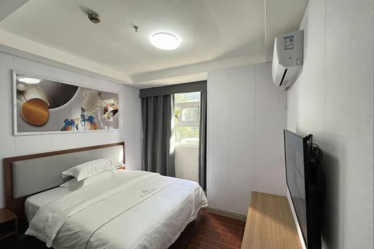 A hotel room with a bed, a TV, a wall-mounted air conditioner, and a window with curtains.