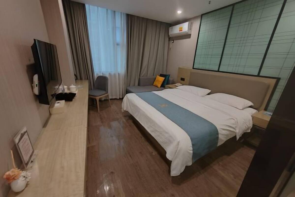 A hotel room with a bed, a desk, a chair, a TV, and an air conditioning unit.
