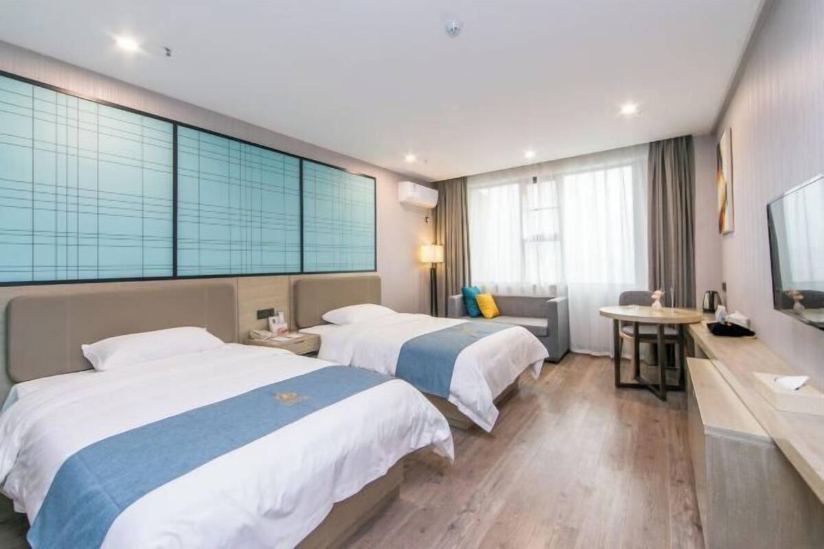 A hotel room with two beds, a desk, a chair, a TV, and a large window with curtains.