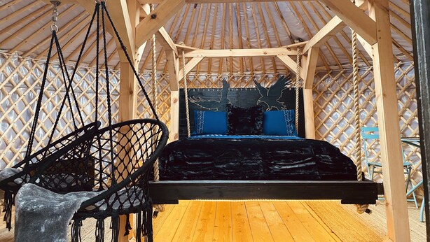 Een houten prieel met een bed dat aan het plafond hangt, een hangstoel en een decoratieve muur.