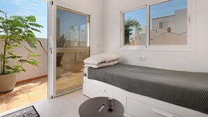 Un petit dormitori amb un llit, una porta corredissa de vidre a un balcó, una planta en test i un comandament a mà a taula.