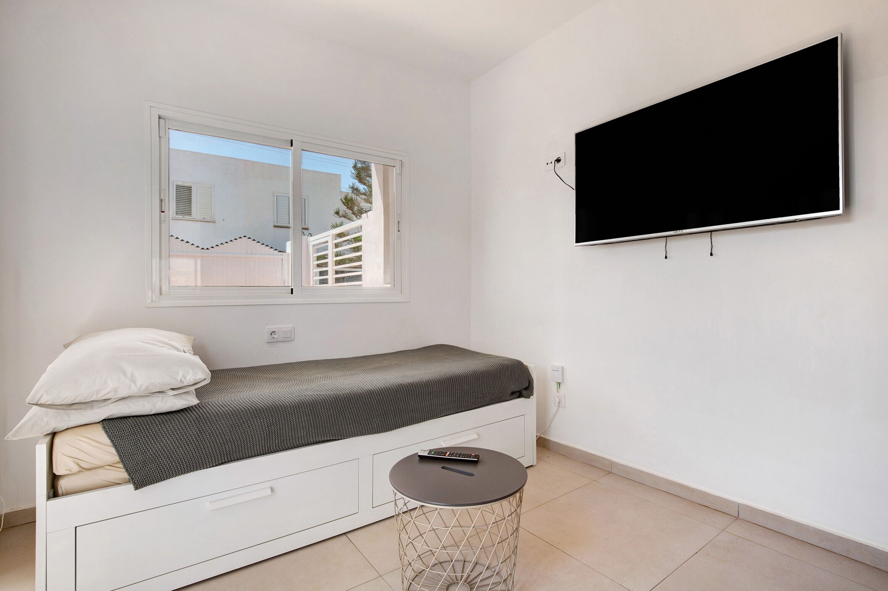 A small room with a bed, a TV mounted on the wall, and a window with a view of a building outside.