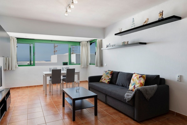 A modern living room with a sofa, coffee table, dining table, chairs, and a view through a green-framed window.