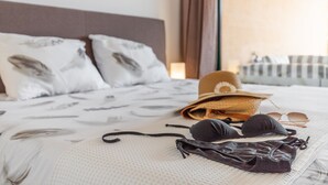 Uma cama com um espalha branco texturizado, duas almofadas estampadas com padrão de pena, um chapéu de palha, óculos de sol e um par de roupa de banho escura sobre a cama.